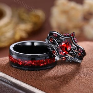 Op de afbeelding: Twee zwarte ringen met rode accenten. Eén ring heeft een grote, rode edelsteen en kleinere rode stenen, met een bloemmotief. De andere ring heeft een band van rode stenen. Beide ringen liggen op een bruin oppervlak.