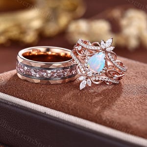 May include: A rose gold engagement ring set. The engagement ring features a pear-shaped opal center stone, surrounded by small diamonds and floral accents. The matching band has a geometric design with pink stones.