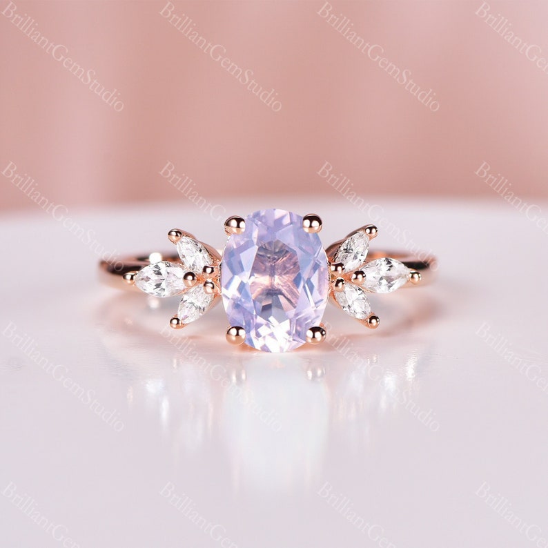 May include: A rose gold engagement ring with a light purple oval-shaped gemstone surrounded by smaller clear gemstones. The ring features a delicate floral design.