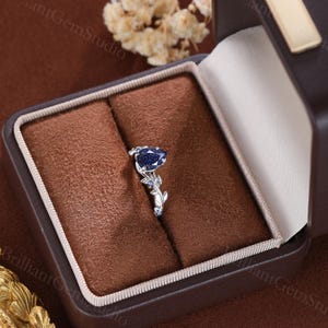 May include: A pear-shaped blue sapphire engagement ring with a delicate floral design. The ring is set in a white gold band and features a cluster of smaller sapphires on the band.