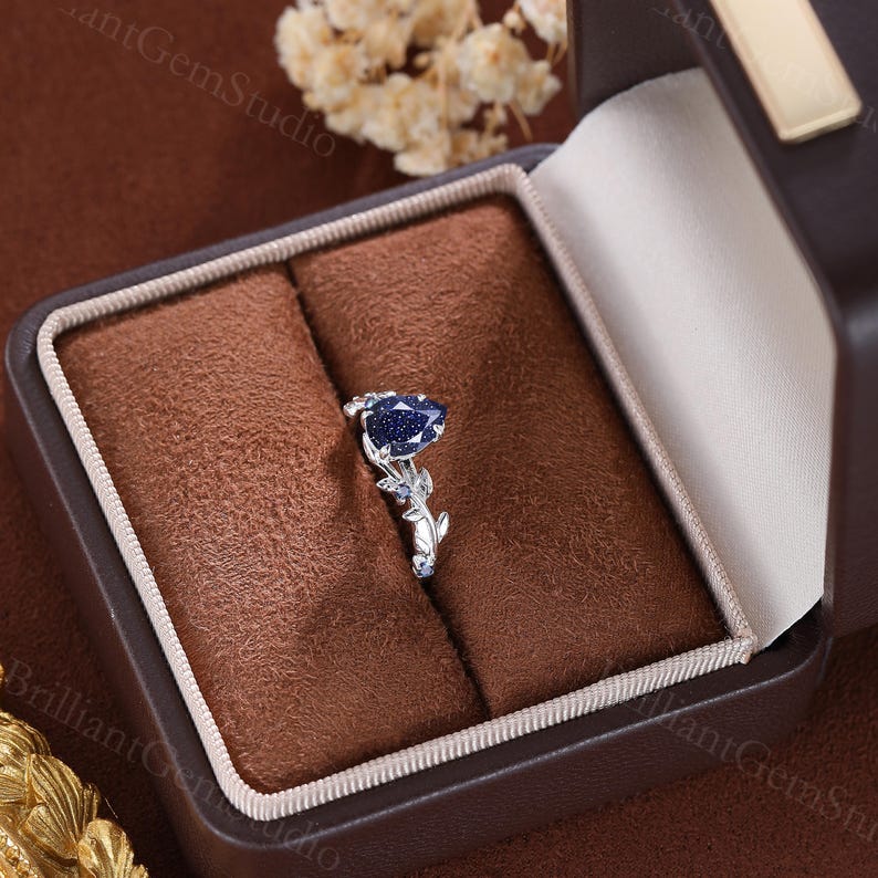 May include: A pear-shaped blue sapphire engagement ring with a delicate floral design. The ring is set in a white gold band and features a cluster of smaller sapphires on the band.