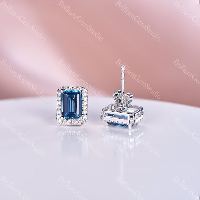 May include: A pair of silver earrings with a blue topaz gemstone set in a square halo of smaller diamonds. The earrings have a post and butterfly back.