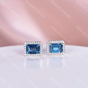 May include: A pair of earrings featuring a rectangular blue topaz gemstone surrounded by a halo of smaller diamonds. The earrings are set in a silver metal.