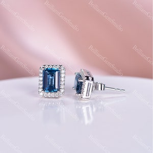 May include: A pair of silver earrings with a blue topaz gemstone set in a square halo of smaller diamonds. The earrings have a post and butterfly back.