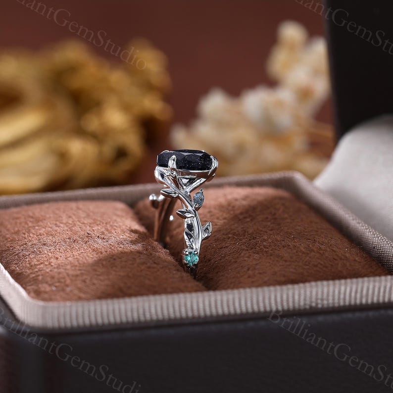 May include: A silver ring with a black oval gemstone and green gemstones on a leafy branch design.