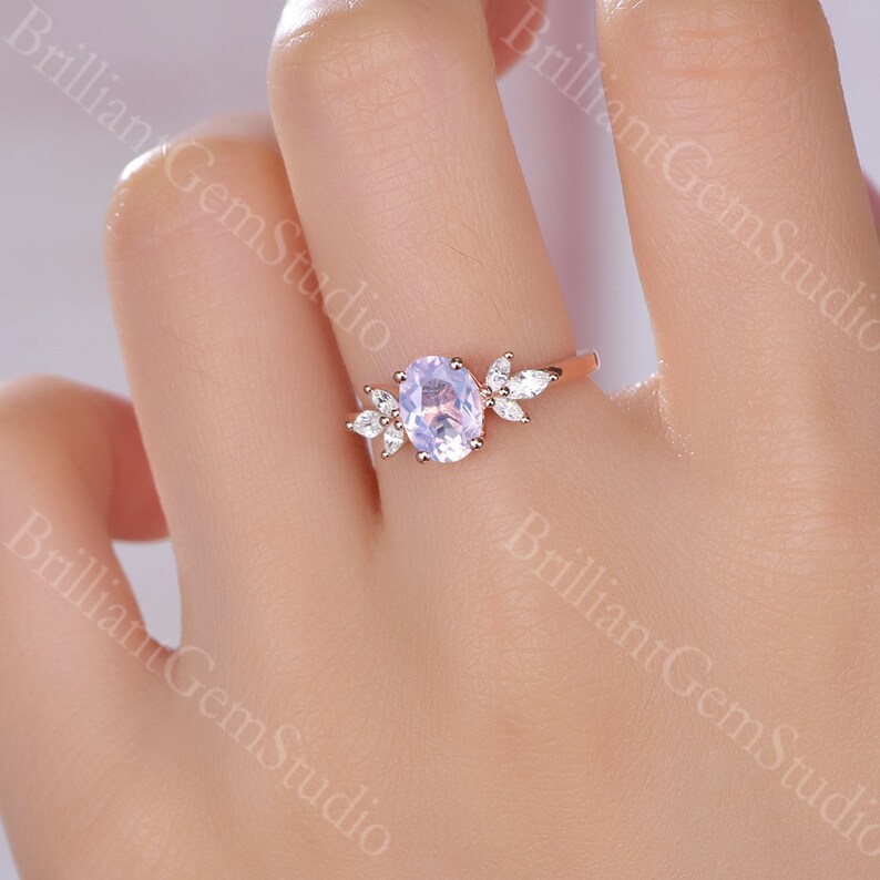 May include: A rose gold engagement ring with a light purple oval-shaped gemstone surrounded by smaller clear gemstones. The ring features a delicate leaf design.