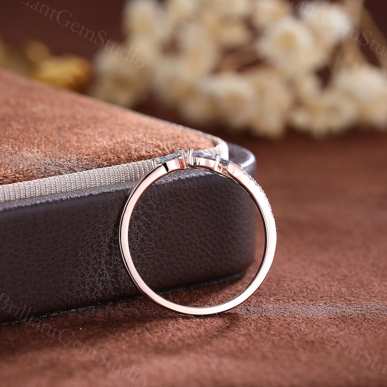 May include: A delicate rose gold ring with a small, clear gemstone set in the center. The band is adorned with smaller, clear gemstones.