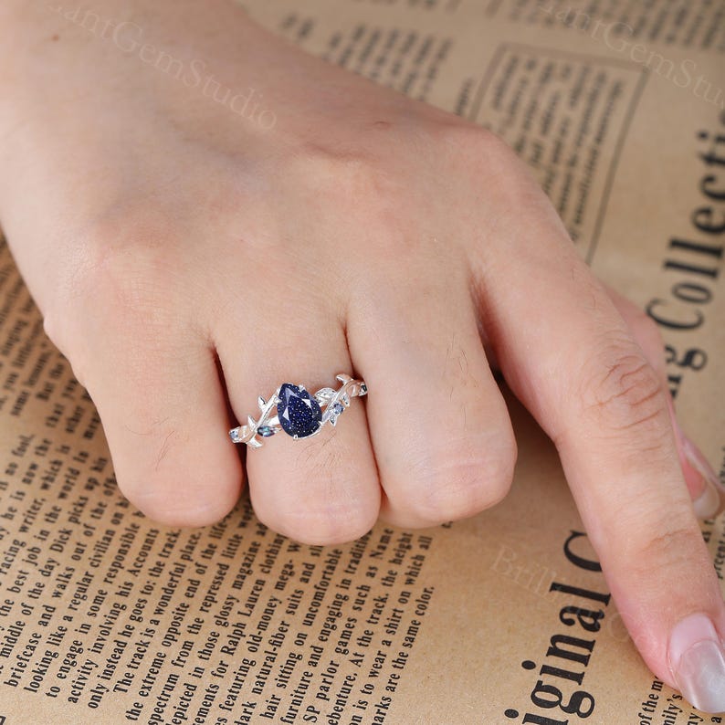 May include: A silver ring with a pear-shaped blue gemstone surrounded by smaller blue gemstones and silver leaves. The ring is on a finger.