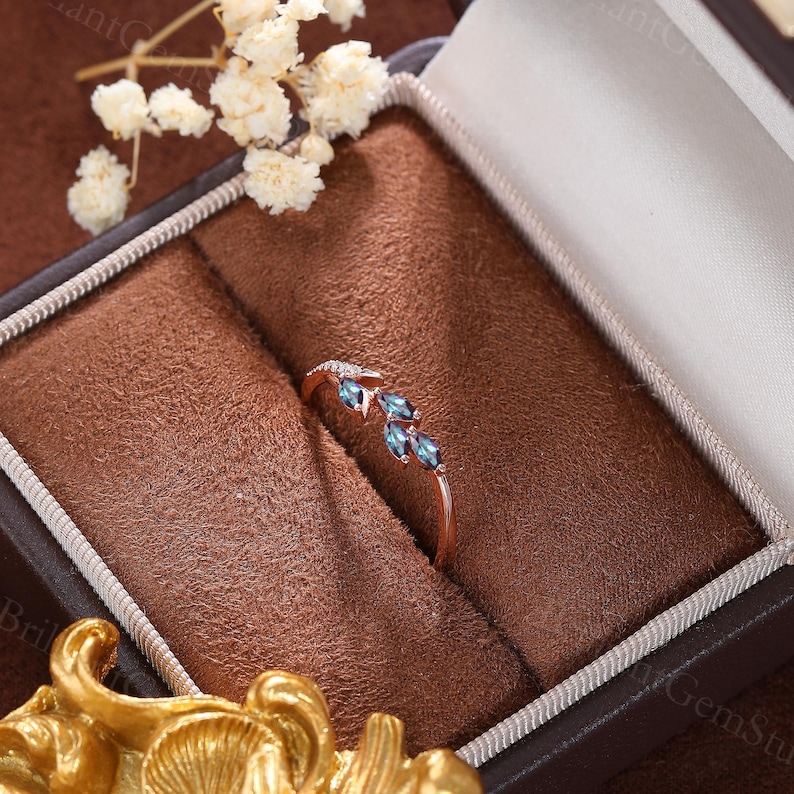 May include: A delicate rose gold ring with three marquise-shaped blue gemstones set in a vine-like design. The ring is displayed in a brown velvet ring box.