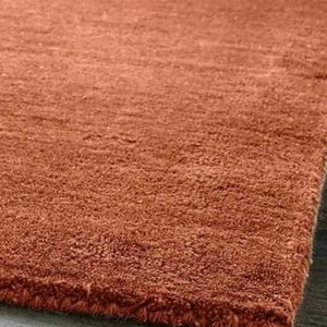 May include: Close-up of a terracotta-colored rug with a textured, plush surface. The rug's fibers create a soft, inviting appearance. The corner of the rug is visible, showing the edge and the dark surface it rests on.