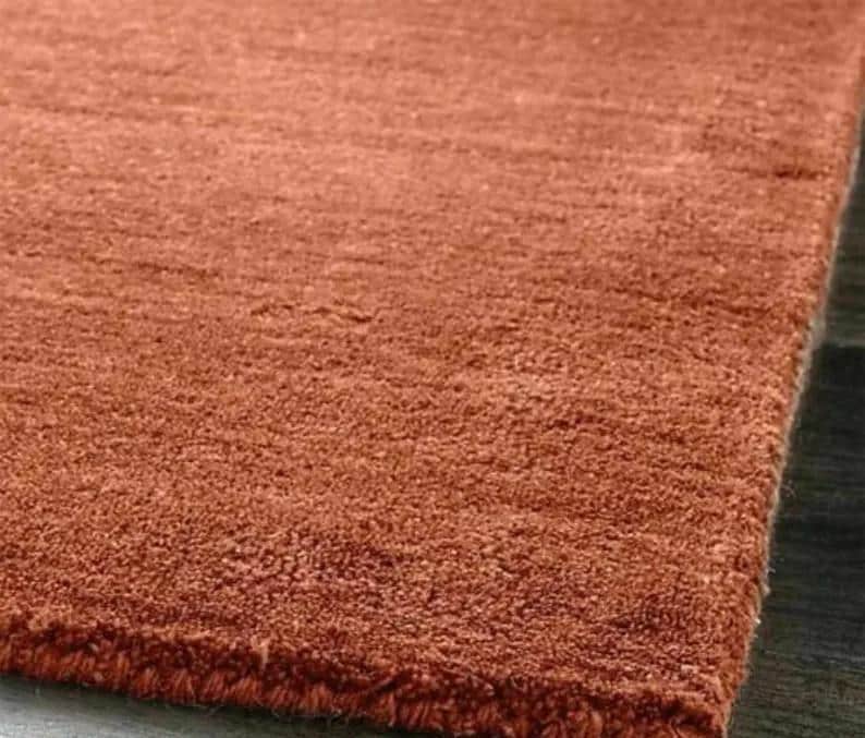 May include: Close-up of a terracotta-colored rug with a textured, plush surface. The rug's fibers create a soft, inviting appearance. The corner of the rug is visible, showing the edge and the dark surface it rests on.