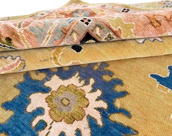 May include: Close-up of a folded rug with a floral pattern. The rug features a combination of colors, including blue, beige, pink, and green. The text "FLORA RUG" is visible on the rug. The rug appears to be made of a soft, woven material.