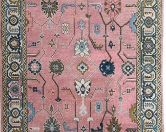 May include: A rectangular pink rug with an intricate floral pattern. The rug features a central field of pink with a repeating design of stylized flowers and geometric shapes in shades of blue, brown, and cream. A detailed border frames the design.