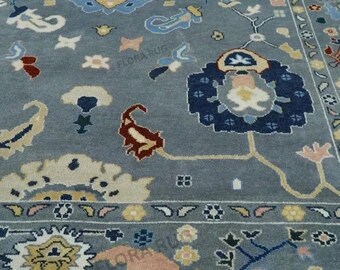 May include: A detailed shot of a gray rug with an intricate floral pattern. The rug features a central design with blue, yellow, and pink elements, surrounded by a border with similar colors and patterns. The rug has a fringe along the bottom edge.