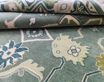 Könnte beinhalten: Nahaufnahme eines aufgerollten, gemusterten Teppichs. Der Teppich weist ein sich wiederholendes Blumenmuster in Grün-, Türkis- und Cremetönen auf. Das komplizierte Muster und die Farbpalette schaffen einen optisch ansprechenden Textilartikel, der sich für die Inneneinrichtung eignet.