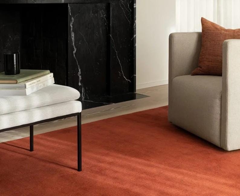May include: A modern living room featuring a burnt orange rug, a white bench with black legs, and a beige armchair with an orange pillow. A black marble fireplace is in the background, creating a warm and inviting atmosphere.