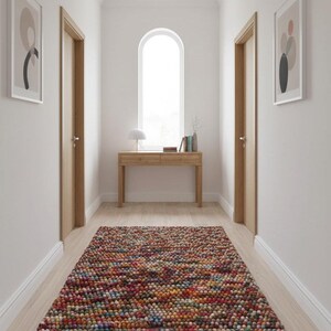 May include: A hallway with a colorful, textured rug. The rug is made of small, round, multi-colored balls. There is a wooden console table with a lamp and books. Two framed abstract art pieces hang on the walls, and two wooden doors are visible.