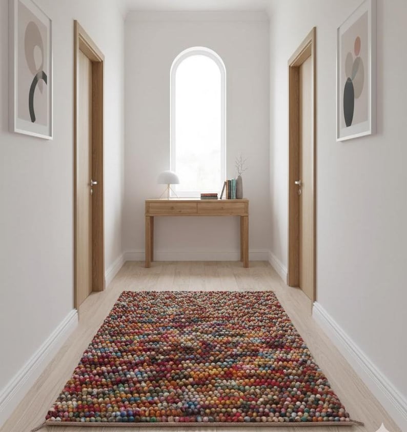 May include: A hallway with a colorful, textured rug. The rug is made of small, round, multi-colored balls. There is a wooden console table with a lamp and books. Two framed abstract art pieces hang on the walls, and two wooden doors are visible.