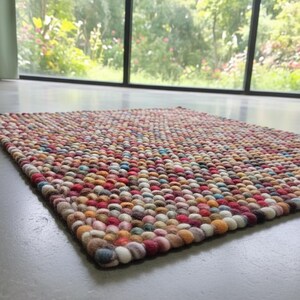 May include: A rectangular rug with a textured surface made of colorful, tightly packed, round felt balls. The rug features a mix of red, orange, blue, and cream-colored balls. The rug is placed on a gray floor, with a window and greenery in the background.