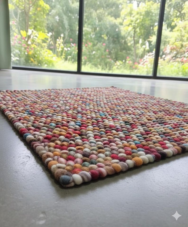 May include: A rectangular rug with a textured surface made of colorful, tightly packed, round felt balls. The rug features a mix of red, orange, blue, and cream-colored balls. The rug is placed on a gray floor, with a window and greenery in the background.
