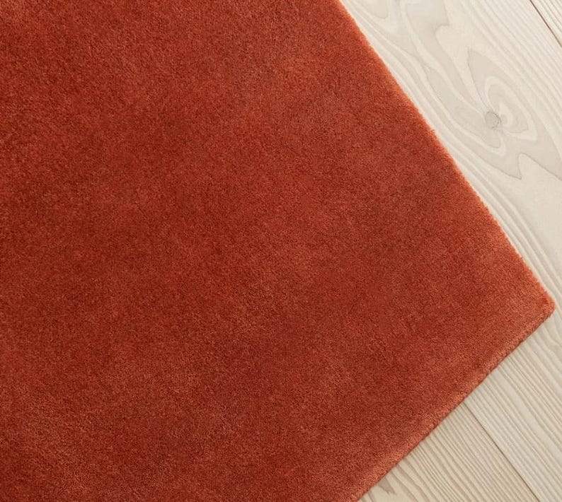 May include: A close-up of a burnt orange rug with a soft, plush texture. The rug is rectangular and fills most of the frame, with a small portion of light-colored wood flooring visible in the corner.