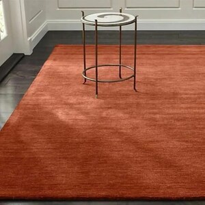 May include: A large, rectangular, rust-colored rug with a textured surface. The rug is placed on a dark wooden floor, with a small side table with a white top and bronze legs in the background. The rug adds warmth and texture to the room.