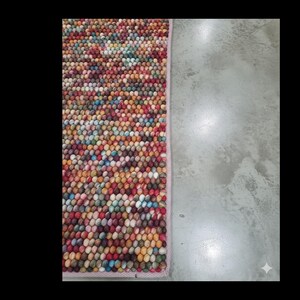 May include: A rectangular rug with a dense, textured surface composed of small, colorful spheres. The rug's color palette includes red, yellow, blue, and green hues. The rug is bordered by a light pink trim.