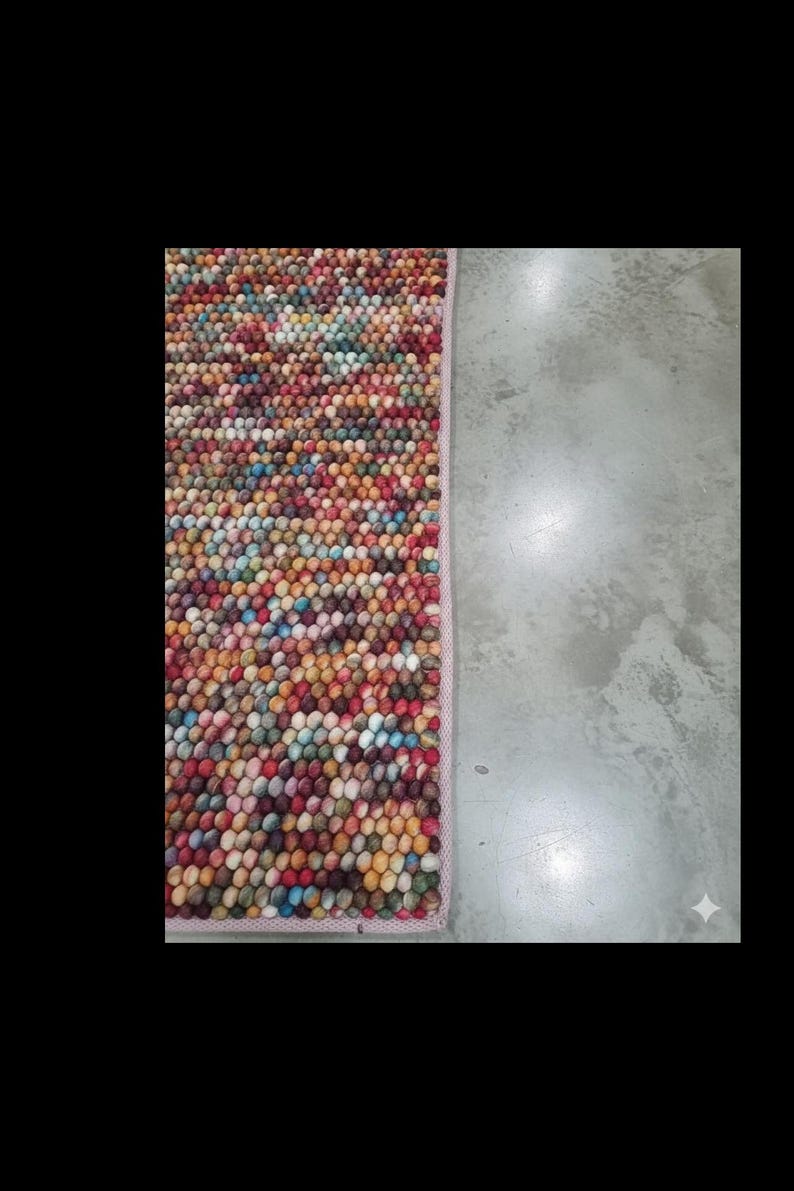 May include: A rectangular rug with a dense, textured surface composed of small, colorful spheres. The rug's color palette includes red, yellow, blue, and green hues. The rug is bordered by a light pink trim.