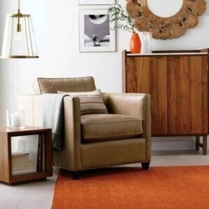 May include: A living room scene featuring a tan leather armchair with decorative pillows and a throw blanket. A wooden side table, a cabinet, and an orange rug complete the space. A pendant light hangs from the ceiling.