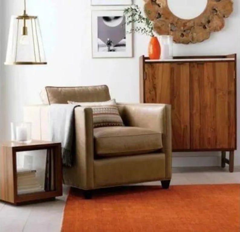May include: A living room scene featuring a tan leather armchair with decorative pillows and a throw blanket. A wooden side table, a cabinet, and an orange rug complete the space. A pendant light hangs from the ceiling.