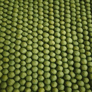 May include: Close-up of a green rug with a textured, bumpy surface. The rug is made of small, round, felted wool balls, creating a unique and tactile design. The color is a muted, earthy green.