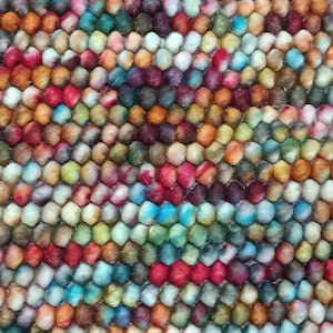 May include: Close-up of a colorful rug with a textured surface. The rug features a pattern of small, round, tightly packed balls in a variety of colors, including red, blue, green, orange, and purple. The rug appears to be made of wool.