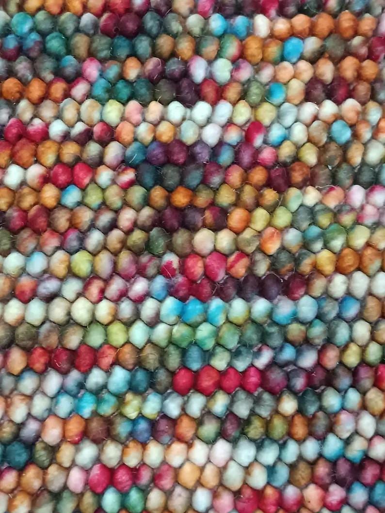 May include: Close-up of a colorful rug with a textured surface. The rug features a pattern of small, round, tightly packed balls in a variety of colors, including red, blue, green, orange, and purple. The rug appears to be made of wool.