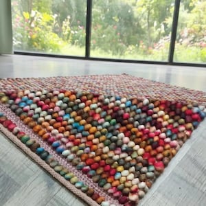 May include: A colorful, handmade rug with a textured surface. The rug is made of small, round, wool balls in a variety of colors, including red, orange, yellow, blue, and green. The rug has a rectangular shape and a woven border.