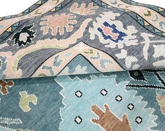 May include: A section of a patterned rug with a folded edge, revealing the underside. The rug features a teal, gray, and pink color palette with floral and geometric designs. The visible edge has a border with a repeating pattern.
