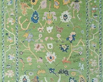 Op de afbeelding: Een groen vloerkleed met een bloemenontwerp. Het vloerkleed heeft een centraal groen veld met kleurrijke bloemen en abstracte patronen in tinten blauw, geel, roze en oranje. De rand is donkergroen met een herhalend geometrisch patroon. De tekst "FLORA RUG" is zichtbaar.