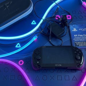 Può includere: Una console di gioco portatile Sony PlayStation Vita nera con custodia protettiva, alimentatore e un set di schede AR Play. La console presenta uno schermo centrale affiancato da tasti direzionali, levette analogiche e pulsanti.