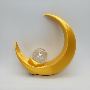 Puede incluir: Una luna creciente dorada con una bola de cristal transparente en su interior. La luna es un objeto impreso en 3D.