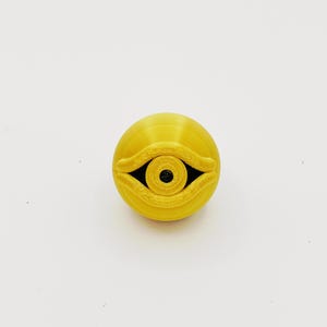 Op de afbeelding: Een felgeel bolvormig object met een verhoogd, gebeeldhouwd oogontwerp op het vooroppervlak. Het oog heeft een prominente ooglidstructuur en een donkere, cirkelvormige pupil in het midden. Het oppervlak toont fijne, horizontale 3D-geprinte lijnen.