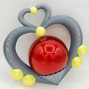 Puede incluir: Un marco gris en forma de corazón con una apariencia texturizada de veta de madera, que presenta una esfera roja grande, lisa y brillante en el centro. El marco está acentuado con varios elementos decorativos circulares de color amarillo pálido.