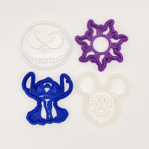May include: Four colorful cookie cutters in the shapes of Jack Skellington, a sun, Stitch, and Mickey Mouse. The Jack Skellington cutter is white, the sun is purple, Stitch is blue, and Mickey Mouse is white. These are likely for baking or crafting.