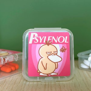 May include: Three clear plastic containers with lids. The container on the left has orange pills inside. The container in the center has a pink label with a cartoon duck on it and the text "PSYLENOL". The container on the right has white pills inside.