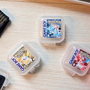 May include: An assortment of video game cartridges and cases, including several labeled "Game Boy" and "Pokemon". A red Nintendo Switch controller is visible on the right. Pens and a keyboard are in the background.