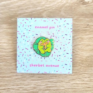 May include: A colorful enamel pin featuring a smiling frog on a lily pad. The pin is set against a light blue background with a confetti pattern. The text "enamel pin" and "sherbet avenue" are visible.