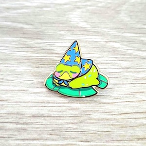 May include: A whimsical enamel pin featuring a sleeping frog wearing a blue wizard hat with yellow stars. The frog rests on a green lily pad with a gold border. The pin has a cute, cartoonish design.