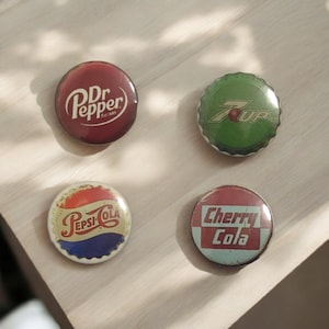 Könnte beinhalten: Vier Vintage-Soda-Flaschenverschlüsse: Dr. Pepper, 7-Up, Pepsi-Cola und Cherry Cola. Die Verschlüsse sind rund mit farbenfrohen Logos und Texten. Der Dr. Pepper-Verschluss ist weinrot, der 7-Up-Verschluss ist grün, der Pepsi-Cola-Verschluss ist rot, weiß und blau, und der Cherry Cola-Verschluss ist rot und weiß.