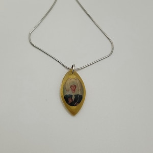 May include: A gold-colored pendant necklace with a silver chain. The pendant is an oval shape with a religious image inside. The image features a person with a halo and dark clothing.