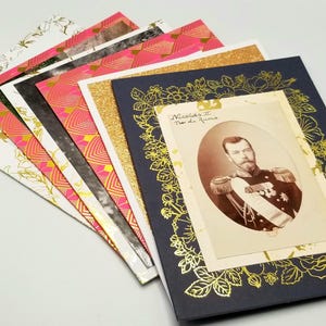 Könnte beinhalten: Eine Sammlung dekorativer Karten mit verschiedenen Mustern und Texturen. Die Karten umfassen florale, geometrische und Glitzerdesigns in Gold, Rosa und Weiß. Eine Karte zeigt ein Vintage-Porträt eines Mannes mit goldenen floralen Akzenten.