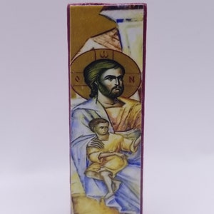 May include: A rectangular, hand-painted religious icon featuring a man with a beard and a child. The man has a gold halo and is wearing a blue and yellow robe. The icon is on a wooden block with a maroon border.