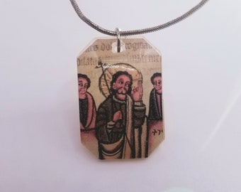 May include: A pendant necklace featuring a rectangular pendant with an illustration of religious figures. The pendant is attached to a silver-colored chain. The illustration has a vintage aesthetic with a light background.
